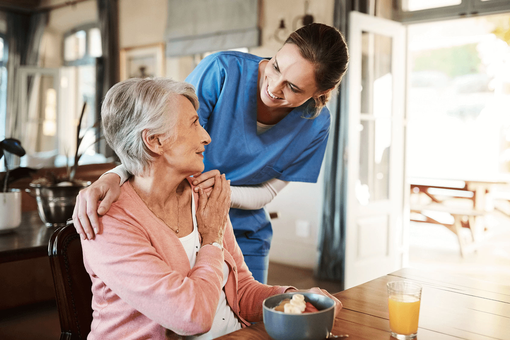 Smiling caregiver in blue scrubs interacting with an elderly woman at a kitchen table with breakfast and juice.